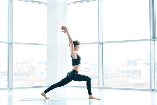 Jeune femme active saine en survêtement noir étirant ses jambes et levant les bras tout en faisant de l'exercice sur le tapis
 - Photo, image