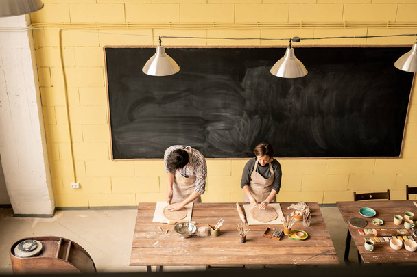 Δύο αυτοαπασχολούμενοι τεχνίτες που εργάζονται με έλασης από πηλό ενώ στέκονται με ξύλινο τραπέζι στο εργαστήριό τους - Φωτογραφία, εικόνα