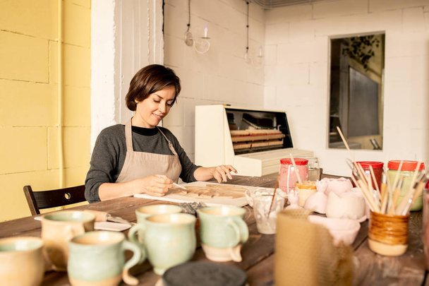 Красивая молодая женщина в рабочей одежде сидит за столом, заканчивая одним из глиняных предметов из новой коллекции в мастерской
 - Фото, изображение