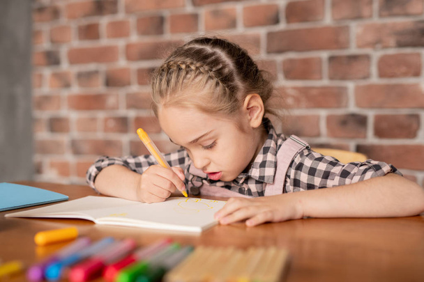 Skoncentrowana kreatywna dziewczyna z plecionych włosów siedzi przy stole i rysunek z filcu końcówki w notebooku w domu - Zdjęcie, obraz