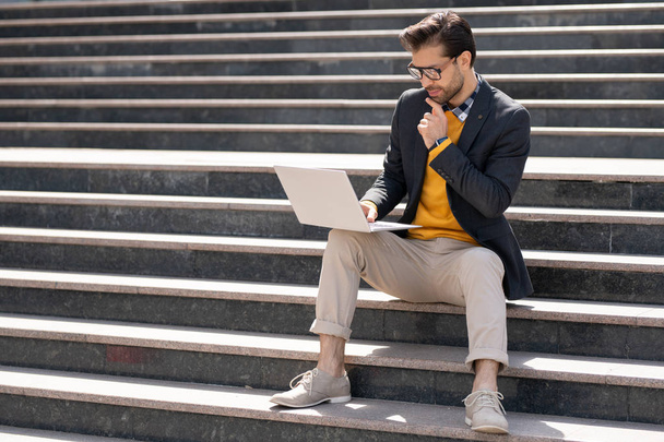 Jovem homem criativo em inteligente casual olhando para a tela do laptop enquanto sentado em escadas em ambiente urbano
 - Foto, Imagem