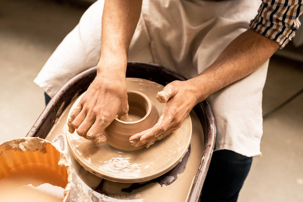 Jovem mestre contemporâneo de cerâmica sentado por roda rotativa enquanto molda panela de argila líquida na oficina
 - Foto, Imagem