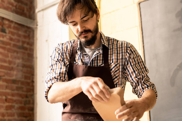 Alfarero barbudo joven en delantal sosteniendo la pieza de trabajo de arcilla mientras va a procesarla en rueda de cerámica durante el trabajo
 - Foto, Imagen