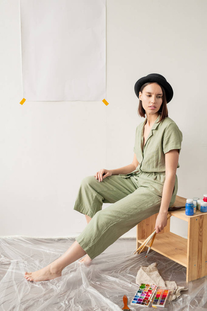 Молодой элегантный художник фримале в повседневной одежде держит в руках кисти, сидя в художественной студии или комнате для рисования
 - Фото, изображение
