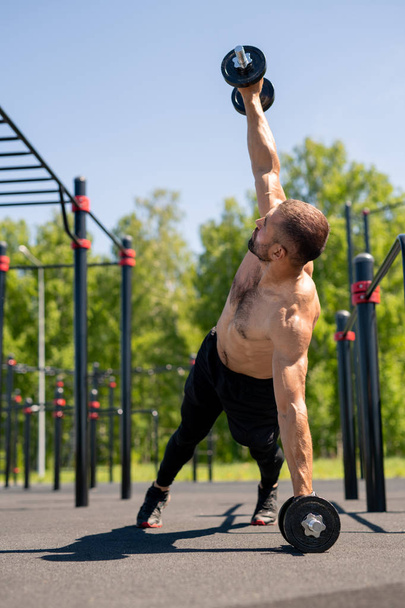 Musclé torse nu gars levant un bras avec haltère lourd tout en s'appuyant sur les jambes et un autre bras pendant l'entraînement en plein air
 - Photo, image