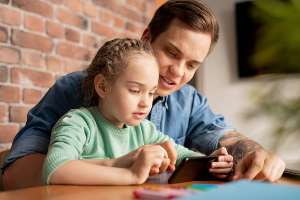Contenido hija curiosa con el pelo trenzado sentado en la mesa y jugando videojuego en el teléfono, mientras que su padre explica cómo completar el nivel
 - Foto, imagen