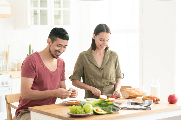 Счастливая молодая женщина режет свежие помидоры на доске, пока ее муж делает овощные сэндвичи
 - Фото, изображение