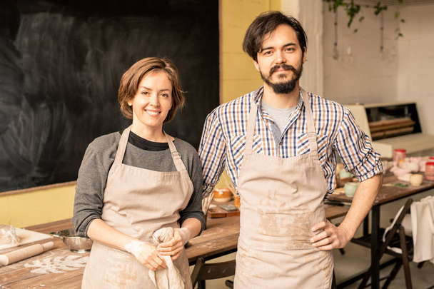 Young man and woman in aprons standing by workplace in front of camera while working together - Photo, Image