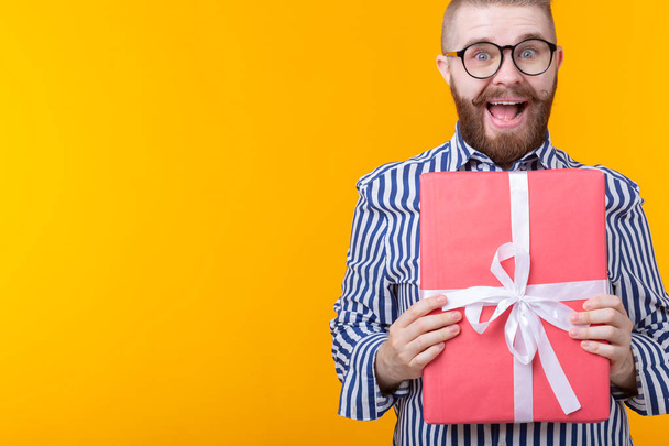 Joyful young hipster guy with a mustache in glasses hugs a red gift box on a yellow background with copy space. The concept of the joy of gifts. - Photo, Image