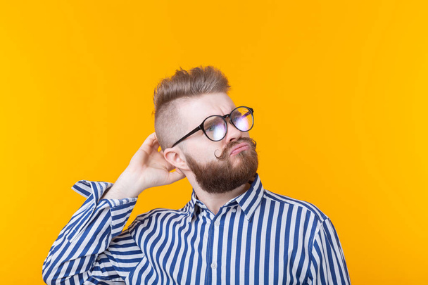 Cute intelligent young man with a beard and with glasses poses on a yellow background and thinks about something. The concept of a difficult choice. - Fotó, kép