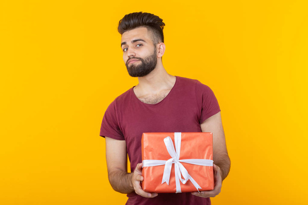 Porträt eines hübschen jungen männlichen Hipsters mit Bart, der auf gelbem Hintergrund posiert und eine rote Geschenkbox in der Hand hält. Konzept von Geschenken und Rabatten. - Foto, Bild