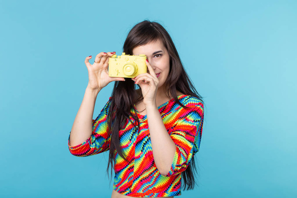 笑顔の若い女性は、コピースペースと青い背景にポーズ黄色のヴィンテージカメラモデルを手に持っています。アマチュアとプロの写真の概念. - 写真・画像