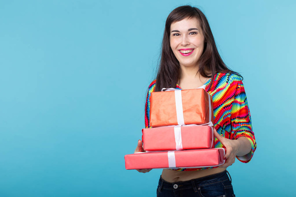 Slim beautiful young brunette girl holding a red gift boxes in her hands while standing against a blue background with copy space. Concept of shares and bonuses. - Photo, image