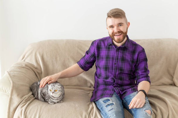 Κομψός νέος άντρας με γένια χίπης χαϊδεύοντας την όμορφη γκρίζα σκωτσέζικη γάτα του κάθεται στον καναπέ. Έννοια της φροντίδας των ζώων. - Φωτογραφία, εικόνα