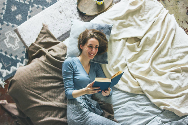 Young woman reading book on bed in loft room, slow life - Fotó, kép