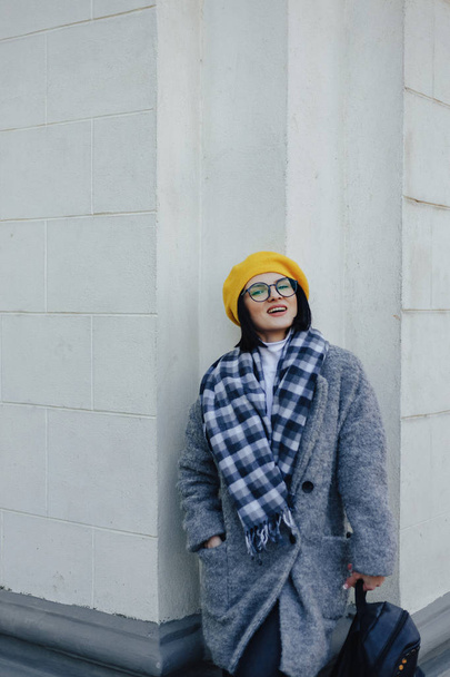 Attractive young girl in glasses in coat and yellow Beret on a simple light background - Valokuva, kuva