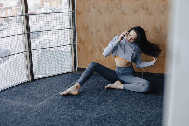 Młoda atrakcyjna dziewczyna fitness siedzi na podłodze w pobliżu okna na tle drewnianej ścianie, spoczywają na zajęciach jogi i rozmawiają na telefon - Zdjęcie, obraz