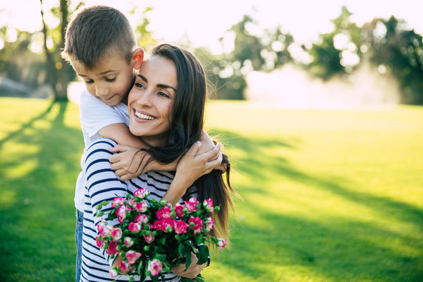 Present on mother's day. Little cute son makes surprise with flowers in hands for his happy and beautiful young mother outdoors in park - Photo, Image