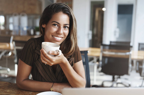 Wellbeing, success, happiness concept. Adorable smiling happy young tanned woman pierced nose hold coffee cup grinning joyfully camera sit cafe table talking friend casually laughing - Fotografie, Obrázek