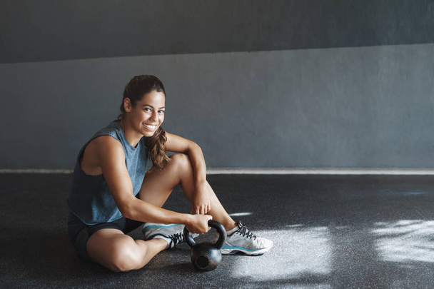 Health, motivation and gym concept. Sportswoman fitness coach take care muscles, rest after crossfit squats, hold kettlebell, sit health club floor, smile camera pleased, sweaty after workout - Fotoğraf, Görsel