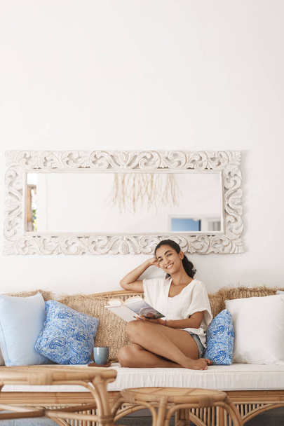 Vertical shot pleased young woman tanned brunette relaxing cozy bright living room lying couch tilt head joyfully smiling camera hold book reading novel drink morning coffee wonderful weekend - Foto, Imagen