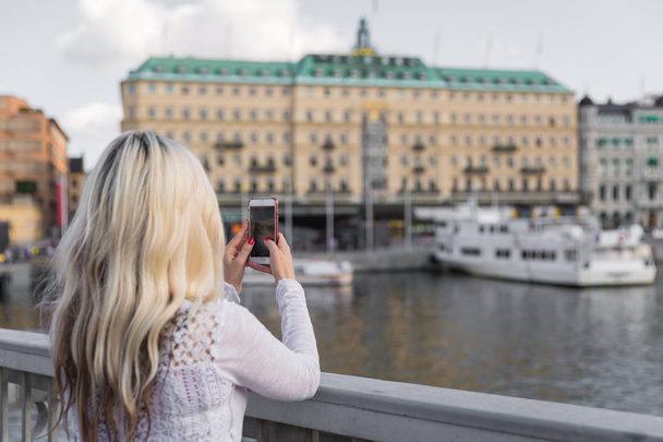 Back view of the young blonde woman who is standing against the background of river and city and doing photo - Foto, Bild