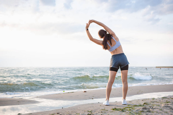 Pretty brunette woman exercising at the sea shore at sunrise listening to the music. - Fotoğraf, Görsel