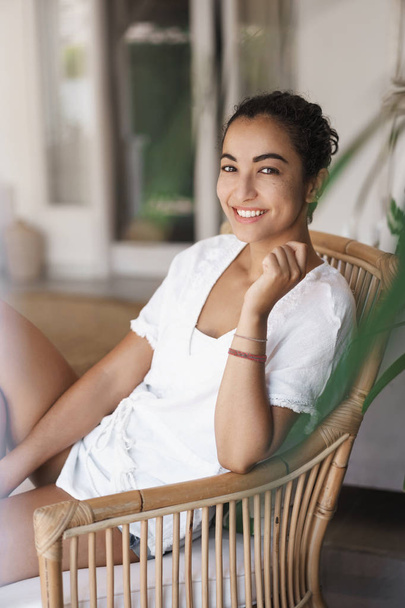 Vertical shot charming smiling tanned brunette woman in white t-shirt sitting rattand armchair, grinning toothy camera joyfully, enjoy summertime vacation, have lazy weekend on home patio - Fotoğraf, Görsel