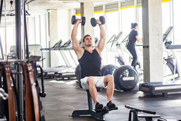 young adult muscular built handsome athlete sitting on bench and holding two dumbbell with raised arms while swinging biceps and working out in gym  - Photo, image