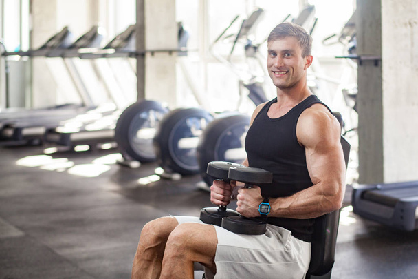 side view of young adult muscular built handsome athlete sitting on weightlifting machine and holding two dumbbell on knees while working out in gym  - Valokuva, kuva