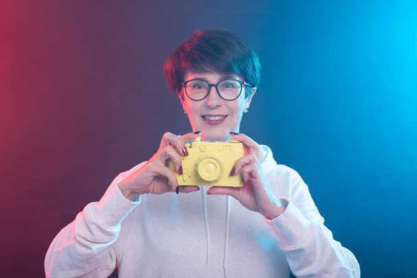 Photographer, art and hobbies concept - Woman 50 years old hold in hand yellow vintage camera on colourful background - Photo, Image