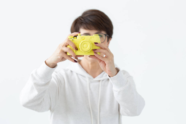 Positive Frau mittleren Alters mit Brille hält eine alte gelbe Kamera in der Hand, die auf weißem Hintergrund posiert. Konzept der Fotoliebhaber und Hobbys - Foto, Bild