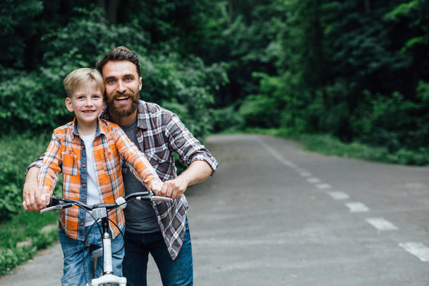 Porträt eines lächelnden Vaters und seines Sohnes, die gemeinsam im grünen Park Spaß haben und dem Sohn beibringen, wie man Fahrrad fährt. Kopierraum für Text. Erlebnisfreizeitkonzept. - Foto, Bild