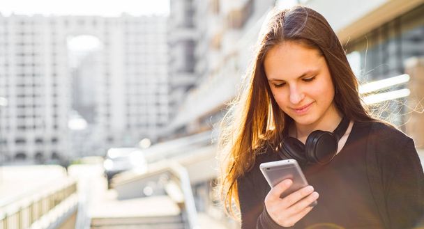 Портрет юной брюнетки-подростка с длинными волосами. девушка на город в черном платье глядя на смартфон на улице в солнечный день
 - Фото, изображение