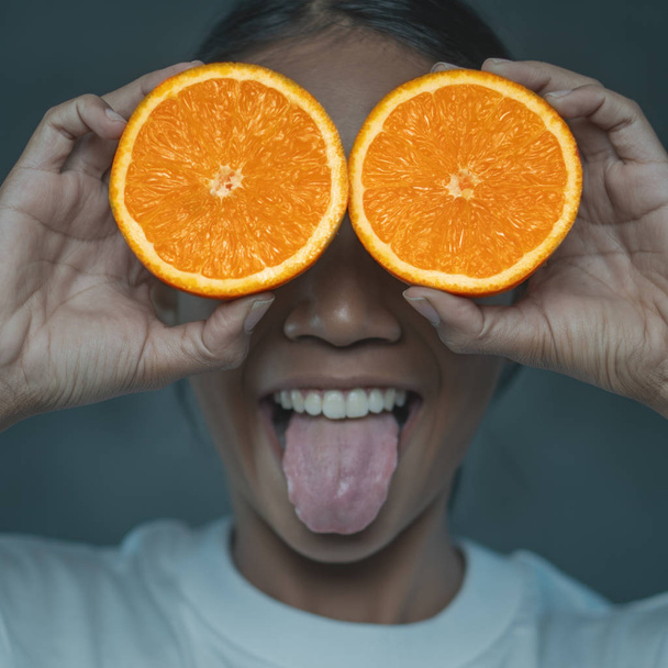 Drôle de portrait de jeune femme montrant la langue tout en couvrant ses yeux avec des moitiés d'orange isolé sur fond de béton
 - Photo, image