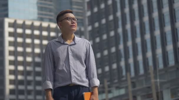 Portré csinos ázsiai üzletember kezében egy könyvet állva körülnézni egy városi város. Lassú-mozgás alacsony szögben lő. - Felvétel, videó