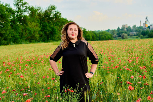 Συν το μέγεθος της γυναίκας με ένα μαύρο φόρεμα σε ένα χωράφι με πράσινο σιτάρι και άγριες παπαρούνες. Υπέρβαρη χοντρή γυναίκα. - Φωτογραφία, εικόνα
