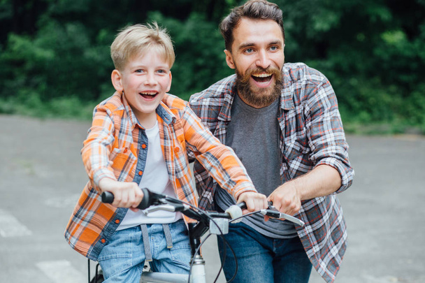Porträt eines lächelnden Vaters und seines Sohnes, die gemeinsam im grünen Park Spaß haben und dem Sohn beibringen, wie man Fahrrad fährt. Erlebnisfreizeitkonzept. - Foto, Bild