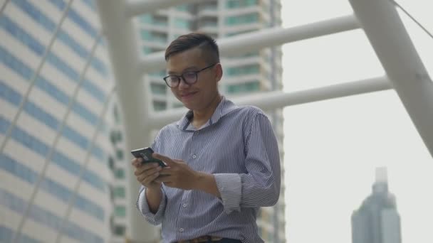 Muotokuva komea aasialainen liikemies hymyilee ja matkapuhelimien avulla lähettää viestin kaupunkilaisessa kaupungissa seisovan ystävän kanssa. Hidastettu kuvaaminen
. - Materiaali, video
