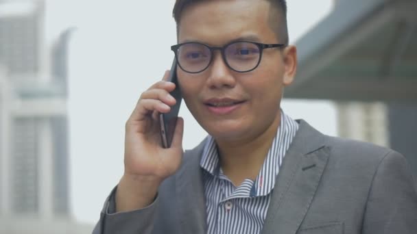 Portrait handsome Asian businessman talking phone with a friend walking on the urban city. An Asian man with glasses uses a phone in the street near a big office building. Slow-motion shoot. - Séquence, vidéo