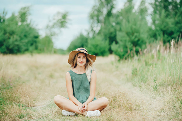 Πορτρέτο του χαμογελαστή κορίτσι στο καπέλο κάθεται σε Λότους ύπαιθρο. - Φωτογραφία, εικόνα