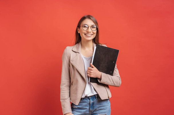 Jonge lachende student of stagiair in brillen staande met een map op de rode achtergrond. - Foto, afbeelding