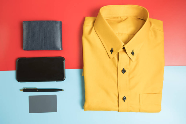 Accesorio para hombre: camisa amarilla, billetera azul, bolígrafo, teléfono móvil y tarjeta de crédito sobre fondo rojo y azul
 - Foto, imagen