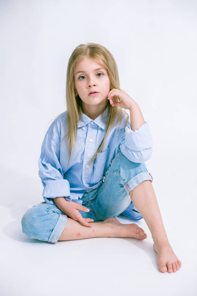 Beautiful fashionable little girl with blond hair in jeans clothes on a white background in studio - Фото, изображение