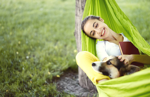 Smiling young woman in green hammock with cute dog Welsh Corgi in a park outdoors. Beautiful happy female in white shirt enjoying good day and hugging a pet. Focus on the dog. - Φωτογραφία, εικόνα