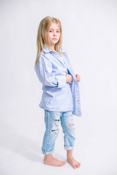 Beautiful fashionable little girl with blond hair in jeans clothes on a white background in studio - Фото, изображение