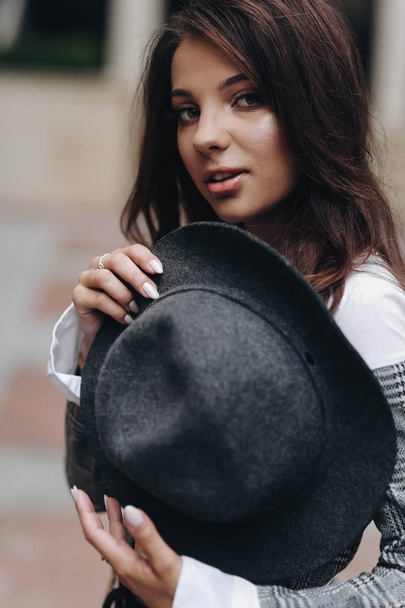 beautiful dark-haired girl in gray jacet hold a hat on her chest  - Zdjęcie, obraz