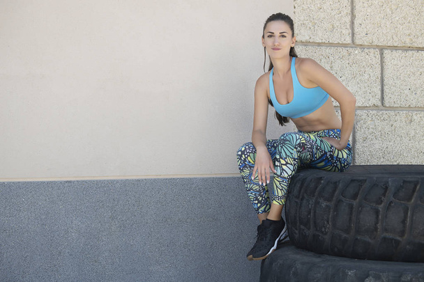 Fit Caucasian woman wearing blue sportswear and sitting on an enormous tractor tire, posing with a pensive, relaxed look after an intense workout routine, healthy lifestyle, sport or fitness concept - Photo, Image