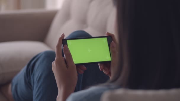 Vonzó fiatal ázsiai nő megtekintése zöld képernyő a mobiltelefon böngészés szociális média míg ülő kanapé a nappaliban otthon. Üzletasszony élvezi néz videókat a smartphone. Slow-Motion lövés. - Felvétel, videó