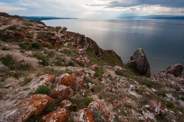 Каміння з червоним мохом на скелях озера Байкал на острові Ольхон. Між камінням зелена трава. Гори позаду озера. Хмари в небі. Сонячні промені сяють крізь хмари. - Фото, зображення